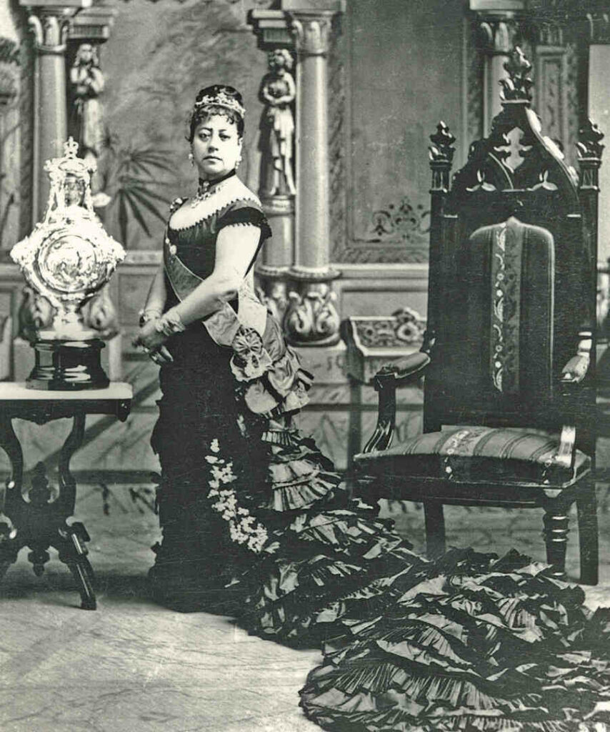Queen Emma of Hawaii in long gown with train standing next to Baptismal urn from Queen Victoria